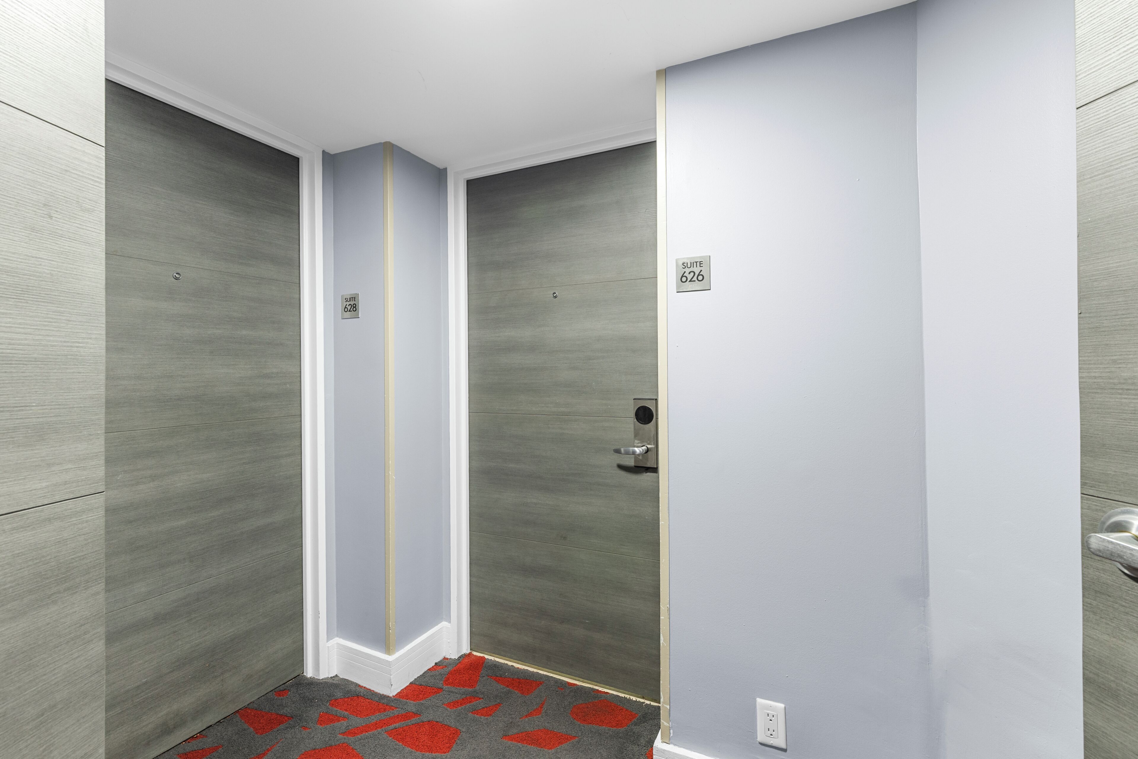 A modern elevator with a grey floor, a red and grey patterned carpet, and a door with a number plate indicating 'SUITE 626'.
