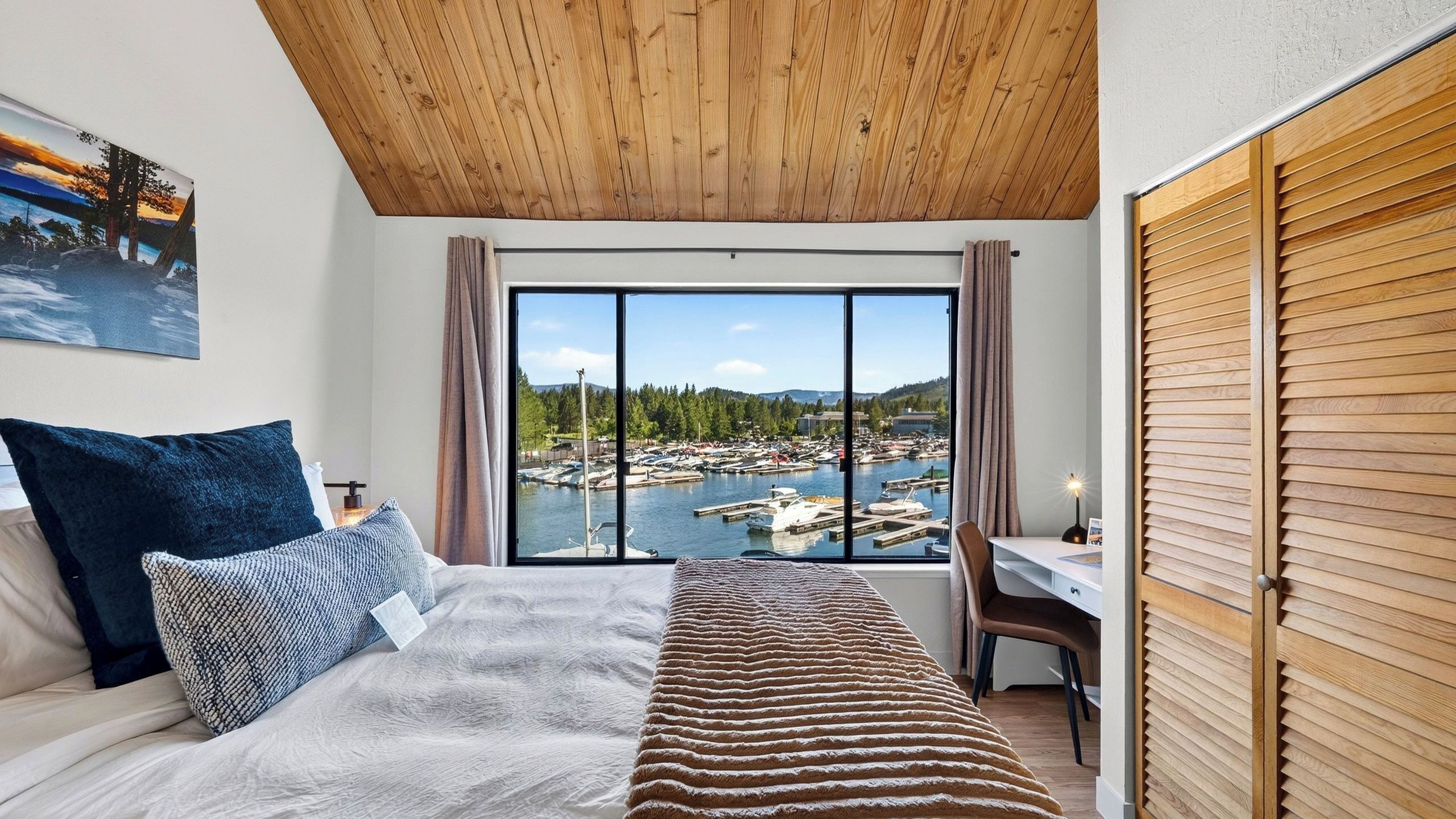 A bedroom with a bed, a desk, a chair, a window with a view of a marina, and a wooden ceiling.