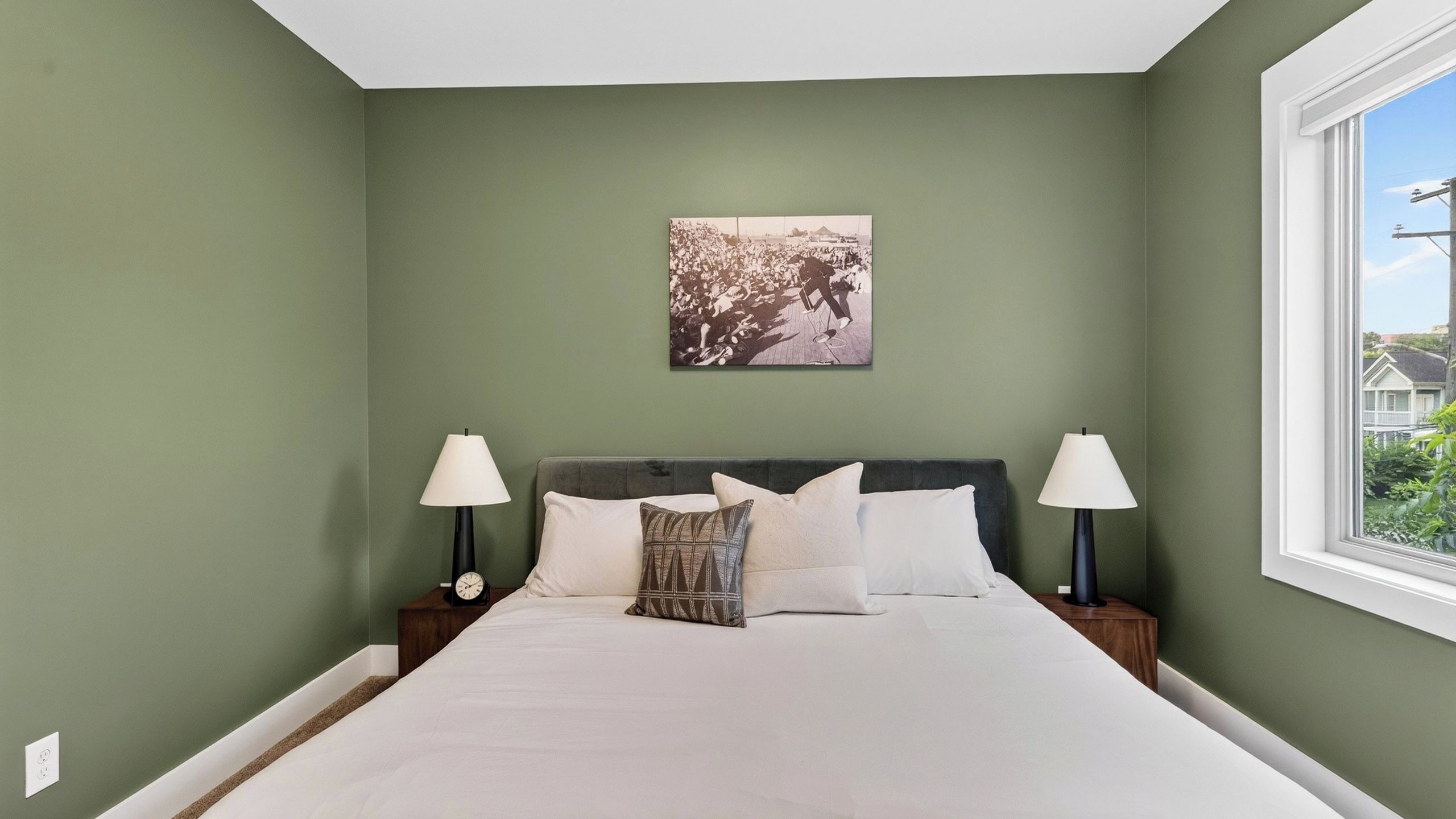A bedroom with a bed, two bedside tables with lamps, a wall clock, and a picture on the wall.