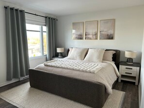 A bedroom with a large bed, two bedside tables, a window with curtains, and three wall-mounted paintings. - Stylish 4-Bedroom Home (Gatineau)
