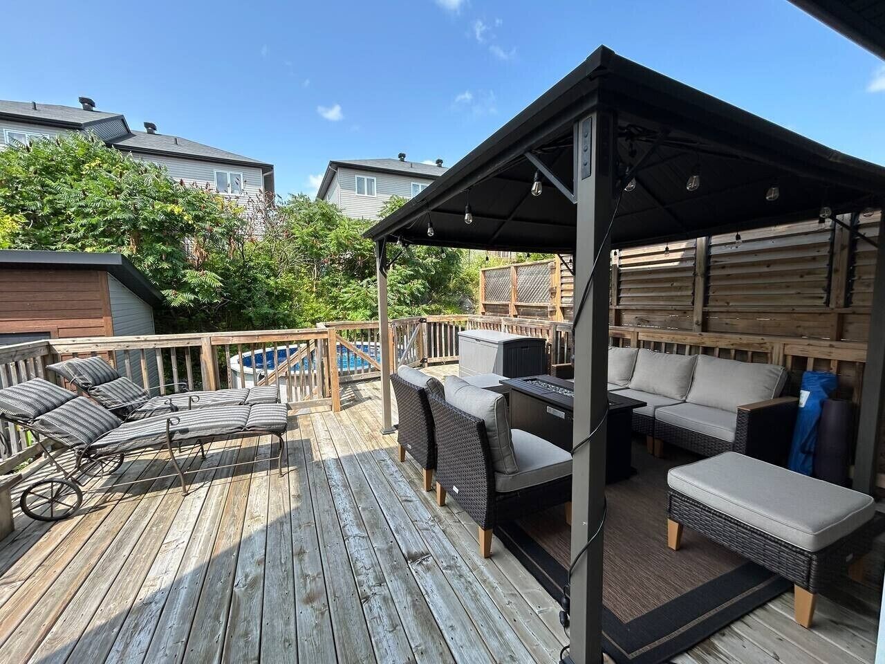 Eine Holzterrasse mit Gartenmöbeln, darunter Liegestühle und eine Sitzecke, unter einem überdachten Bereich.