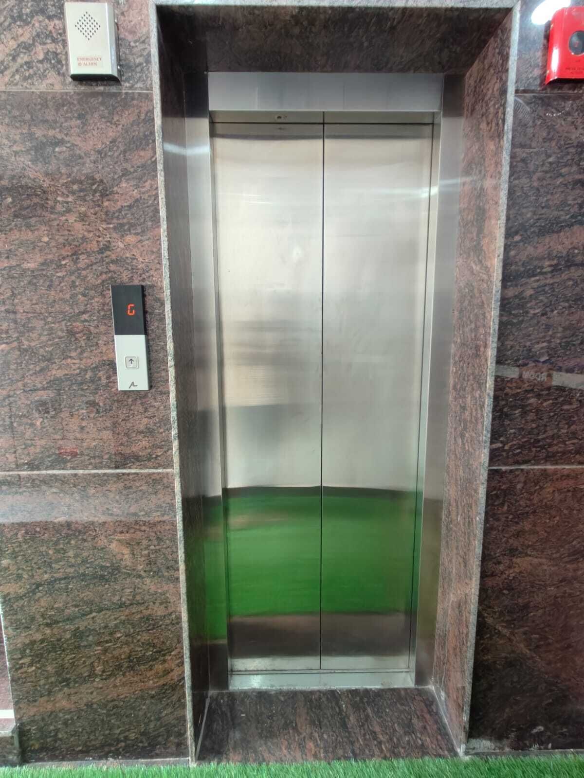 An elevator with a metallic interior, a green floor, and a control panel with an emergency alarm button.