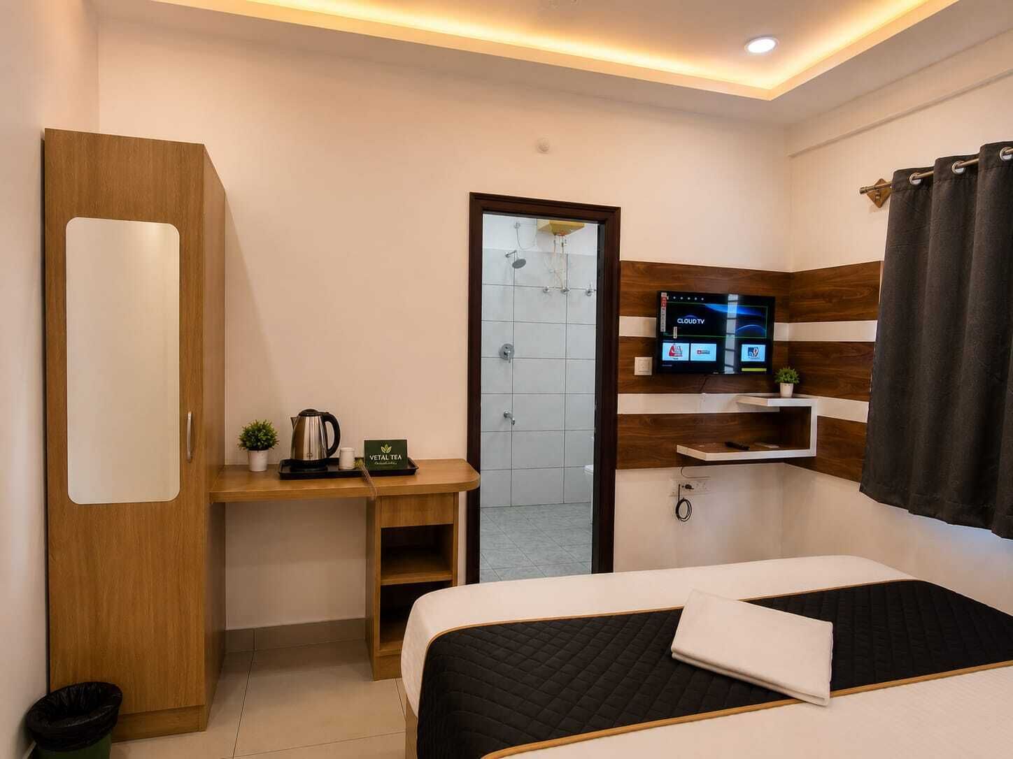 A hotel room with a bed, a desk with a kettle, a TV, and a shower area.