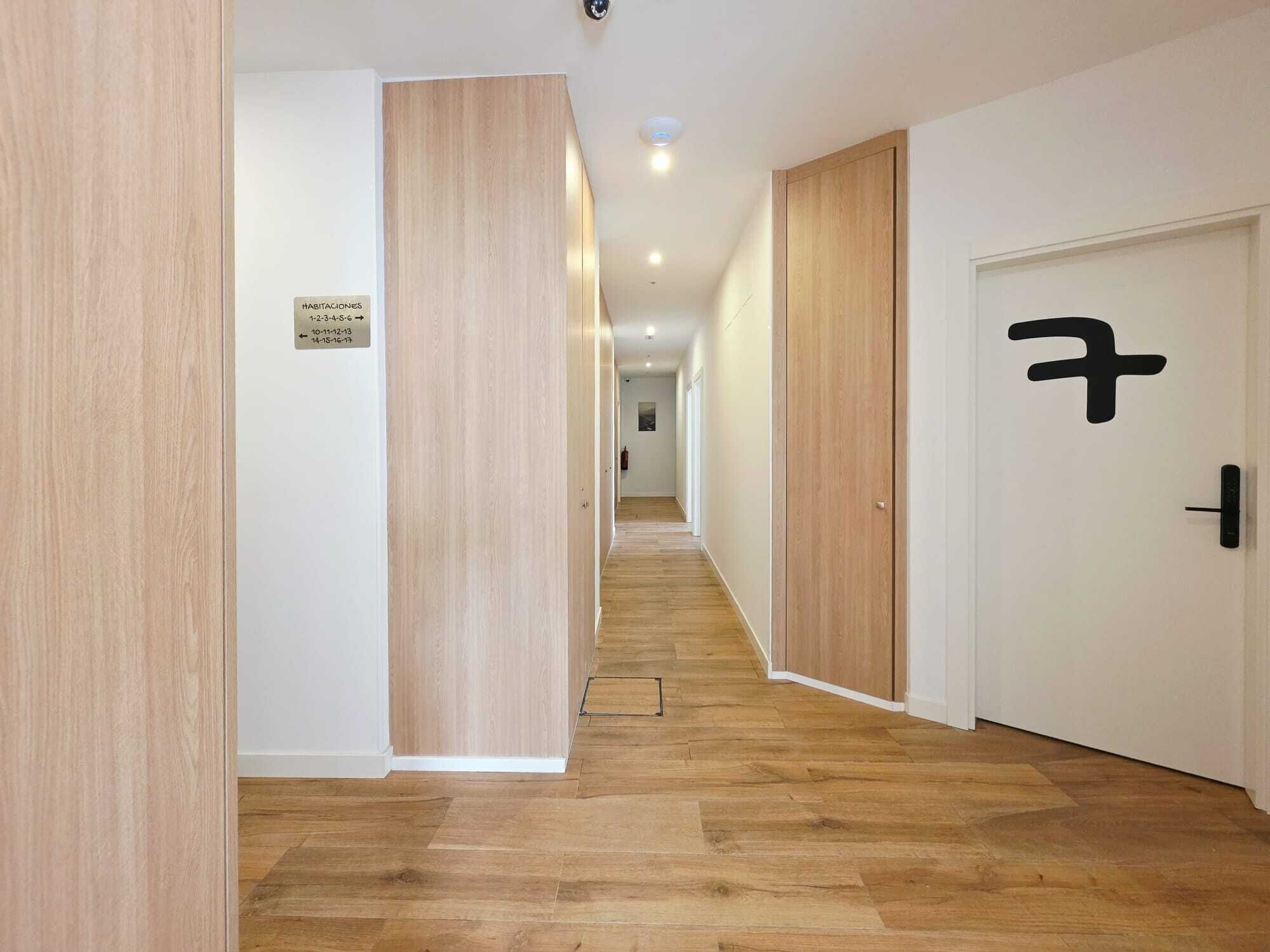 Un pasillo con piso de madera, paredes blancas y puertas con manijas negras. Un cartel indica la disponibilidad de habitaciones.