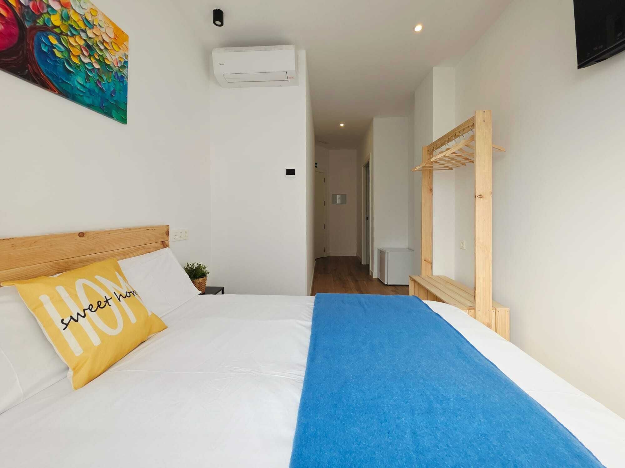 Una camera d'albergo con un letto, un condizionatore d'aria a parete, una testiera in legno, un asciugamano blu e un quadro colorato.