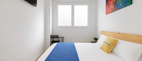 A bedroom with a bed, a TV mounted on the wall, a window, and a painting.