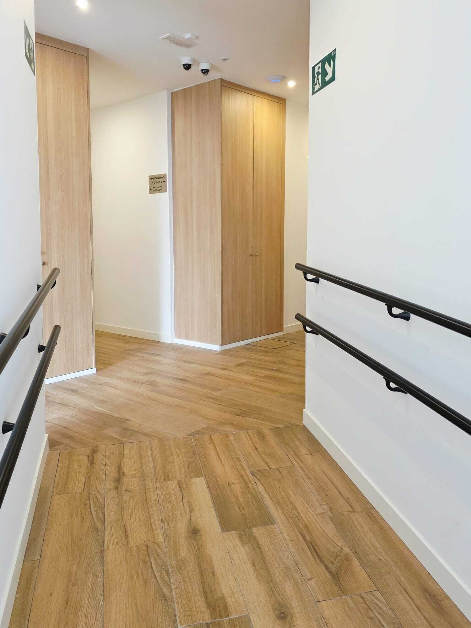 A hallway with wooden flooring, a white wall, and a door with a green exit sign.