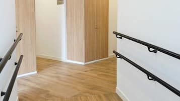 Un pasillo con piso de madera, pared blanca y una puerta con letrero de salida verde.