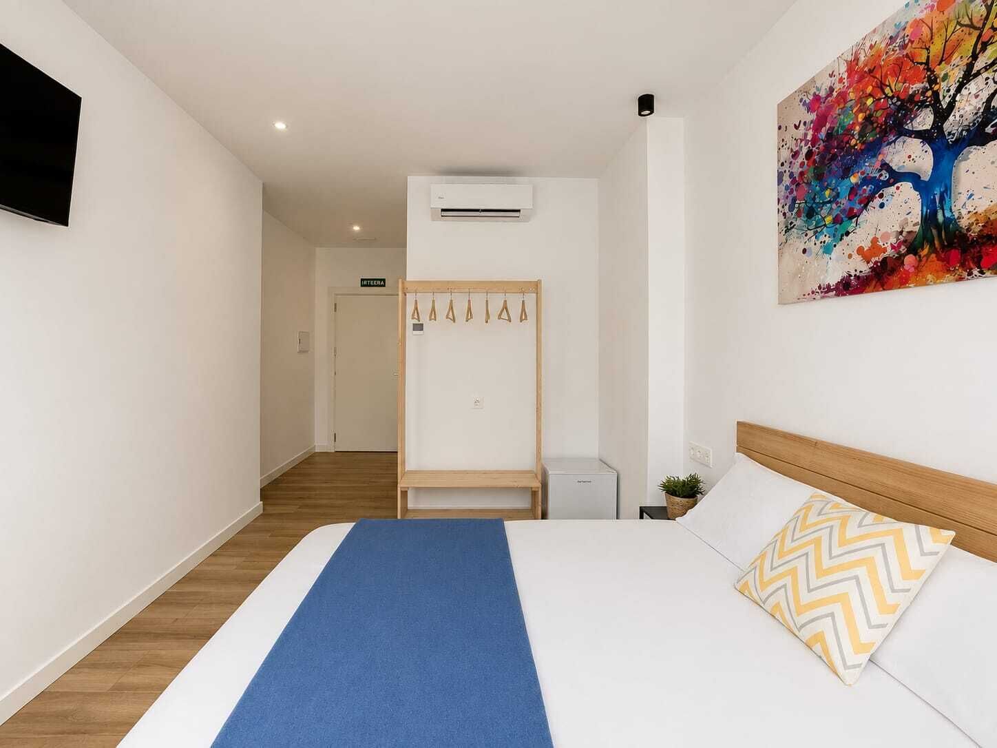 A hotel room with a bed, a TV mounted on the wall, an air conditioning unit, a coat rack, and a colorful painting.