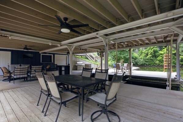 An outdoor covered patio with a table and chairs, a ceiling fan, and a view of a dock and greenery.