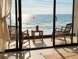A room with a view of the ocean, featuring two wooden chairs, a small table with a cactus, and a glass door. - Luxury Beachfront with King Bed & AC (Tijuana)