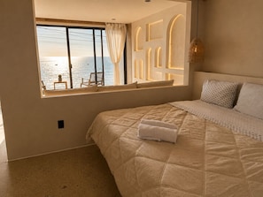 A bedroom with a large bed, a window with a view of the sea, and a small table with a chair outside the window. - Luxury Beachfront with King Bed & AC (Tijuana)