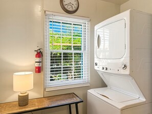A laundry room with a white washer and dryer, a fire extinguisher, a window with blinds, and a small table with a lamp. - Casa de las Flores (Santa Fe)