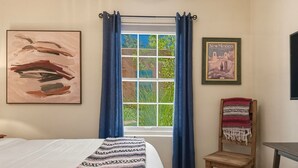 Ein Schlafzimmer mit einem Bett, einem Fenster mit Vorhängen, einem Sessel und zwei gerahmten Bildern an der Wand.