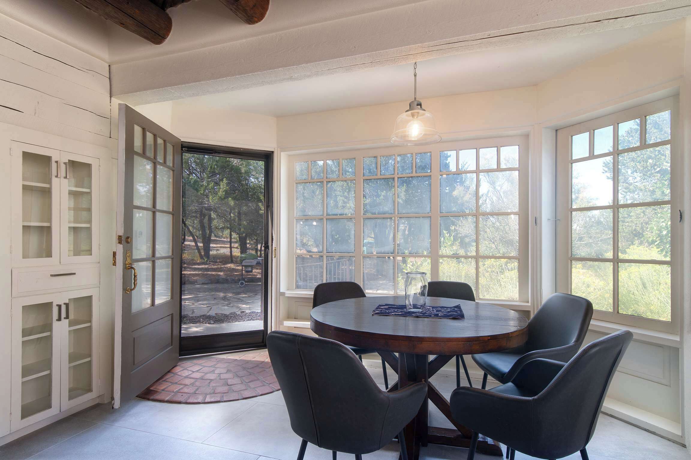 A dining area with a round table, four chairs, a hanging light fixture, and large windows with a view of trees.