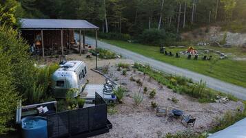 A rustic cabin with a covered outdoor seating area, a camper, and a fire pit in a forested setting.