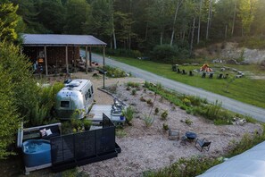 Eine rustikale Holzhütte mit überdachter Außenveranda, einem Wohnmobil und einem Feuerkorb inmitten eines bewaldeten Geländes.