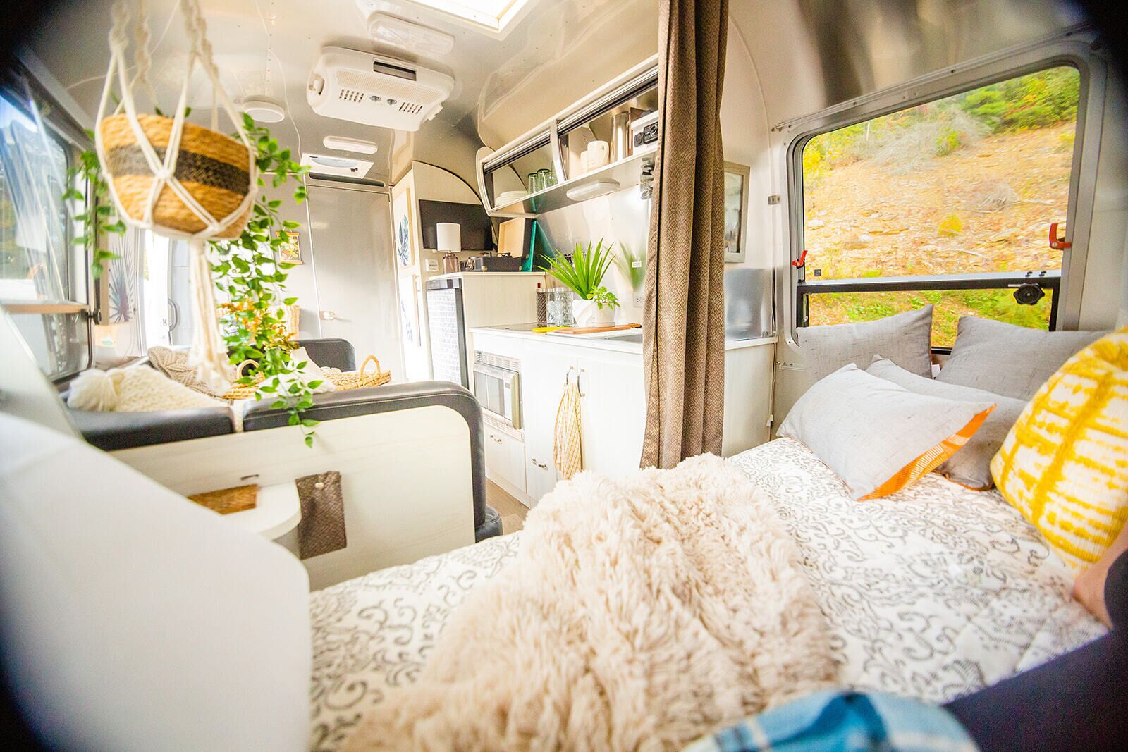 A cozy RV interior with a bed, kitchenette, and a window with a view of a forest.