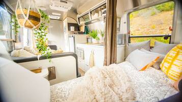 A cozy RV interior with a bed, kitchenette, and a window with a view of a forest.
