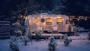 A stationary RV with a covered porch area, surrounded by snow-covered plants and trees.