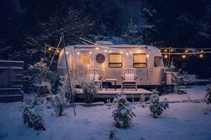 Ein stehender Wohnmobil mit überdachter Veranda, umgeben von schneebedeckten Pflanzen und Bäumen.