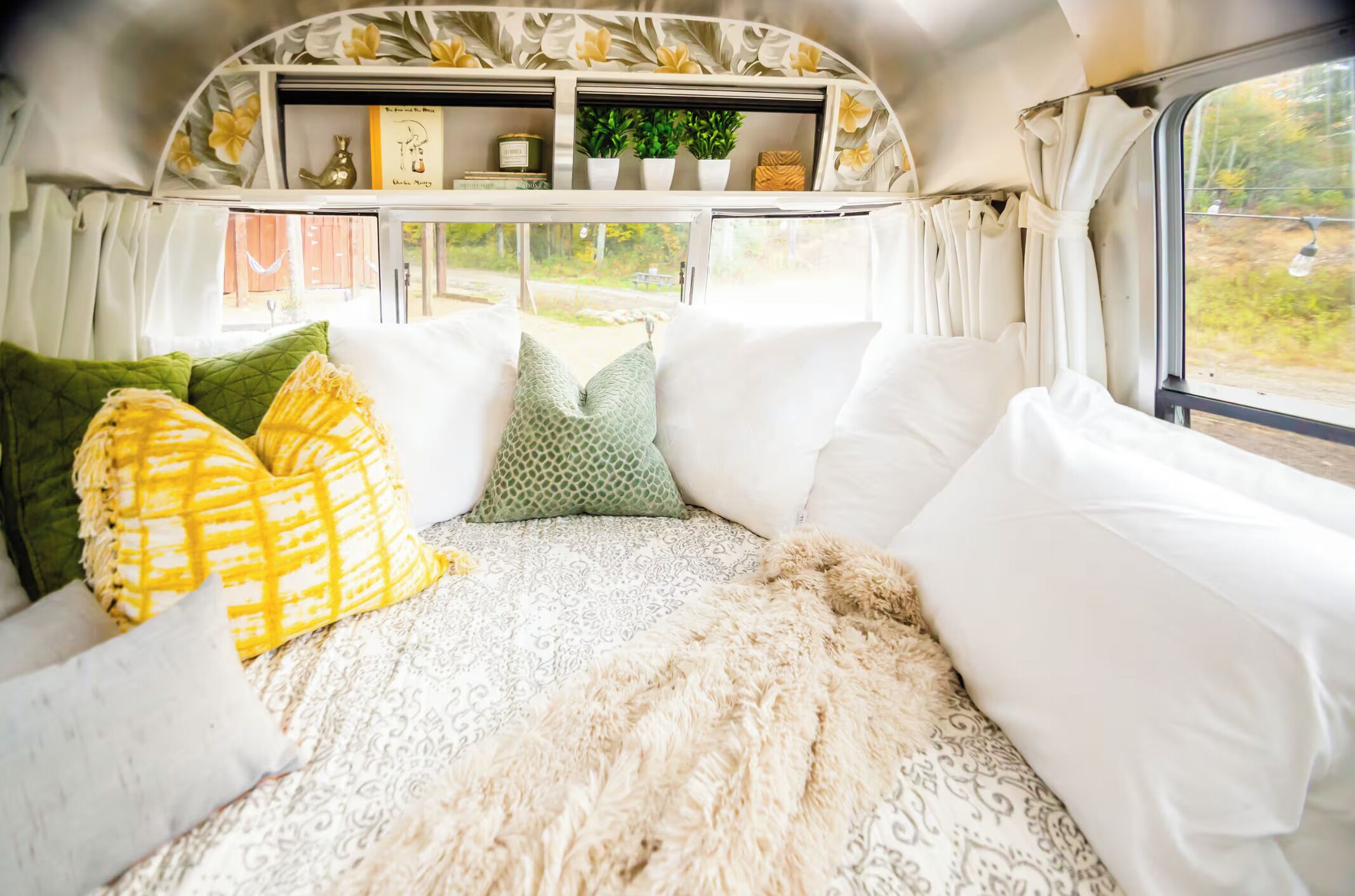 Un intérieur chaleureux avec un lit, des oreillers, un plaid en fourrure et une fenêtre donnant sur un paysage verdoyant.