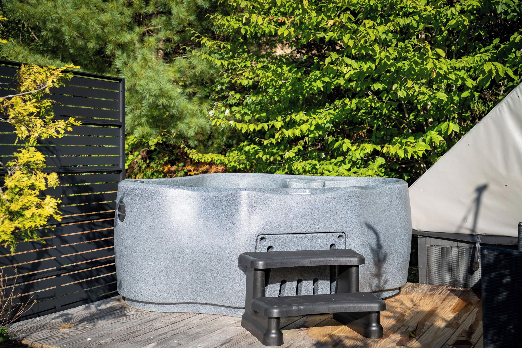 Un jacuzzi en una terraza de madera con cerramiento negro y follaje al fondo.