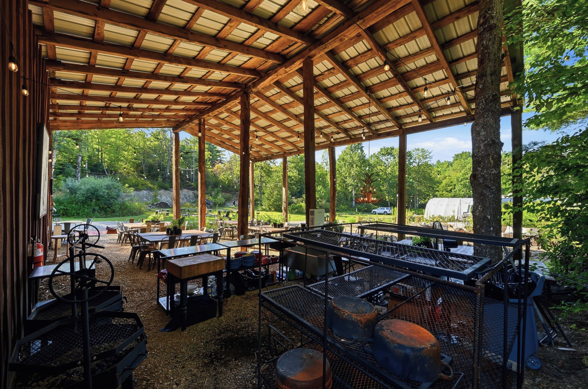 Un patio cubierto al aire libre con mesas y sillas, rodeado de vegetación y un invernadero al fondo.