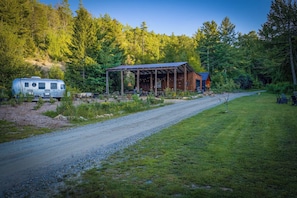 A rustic cabin with a covered porch, a gravel path, and a trailer in a forested area. - Petite Airstream Cottage w/Private Hot Tub + Sauna Access by Blue Ridge Parkway (Newland)