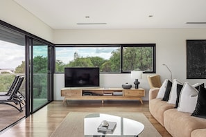 Un moderno salón con televisor grande, muebles de madera y una pared de vidrio con vista a follaje.