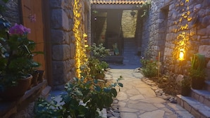 Un chemin de pierre menant à l’entrée d’un bâtiment, bordé de plantes en pot et entouré de murs de pierre.