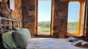 Un lit recouvert d’une couverture à carreaux, avec deux oreillers et des serviettes, adossé à un mur en pierre et doté d’une fenêtre donnant sur un paysage.