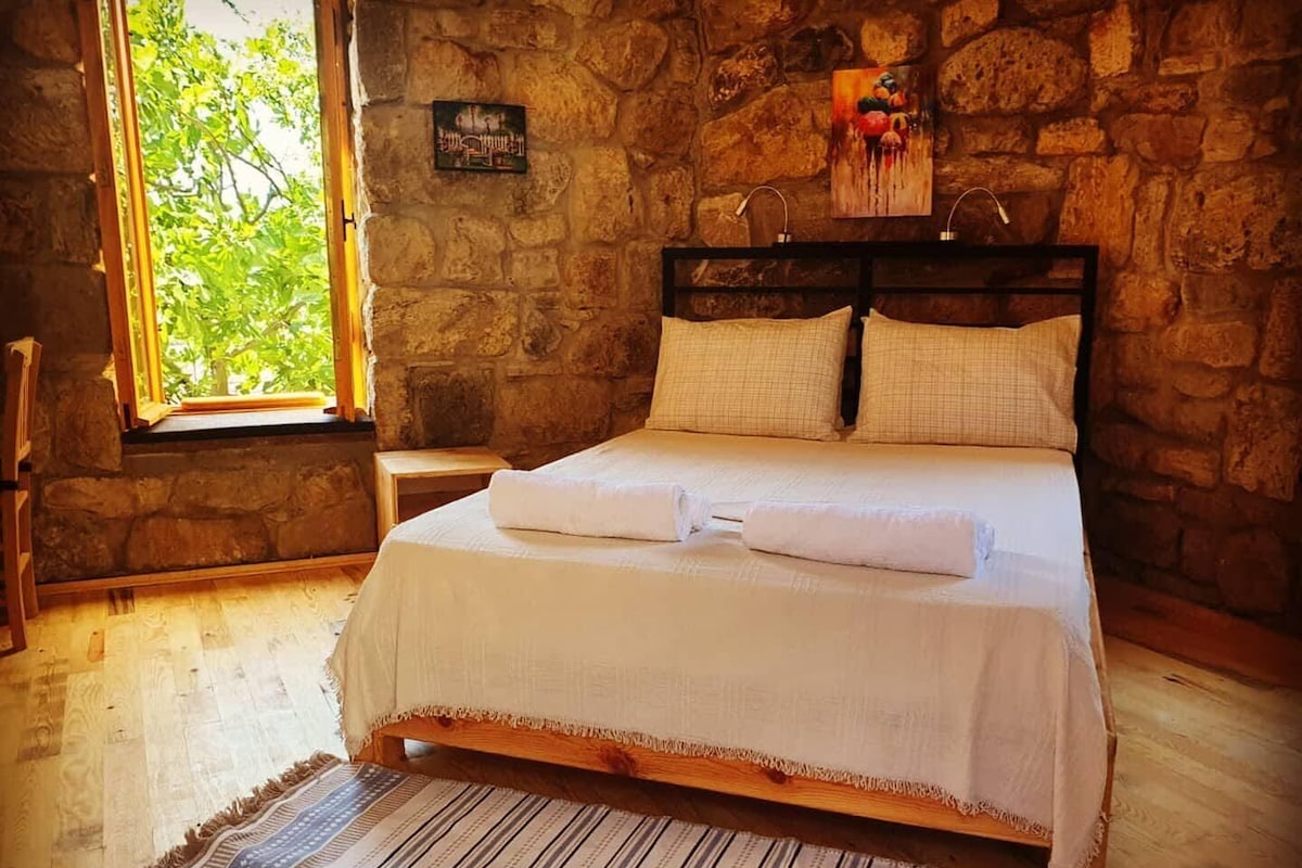 Una habitación con pared de piedra, cama con ropa de cama blanca, ventana con vista a follaje y un cuadro en la pared.