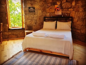 A room with a stone wall, a bed with white bedding, a window with a view of greenery, and a painting on the wall.