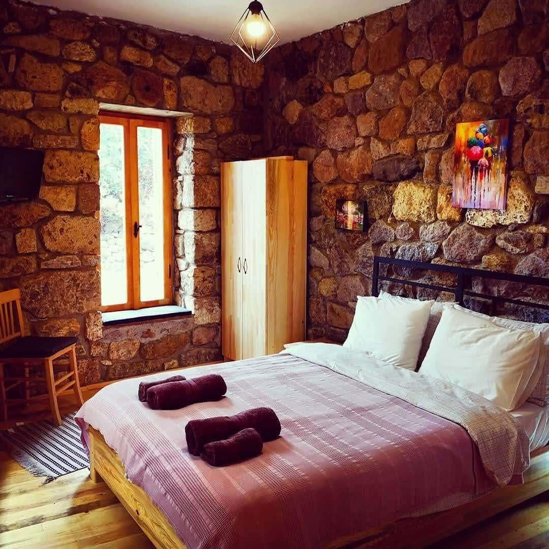 A room with a stone wall, a bed with a pink bedspread, a wooden wardrobe, and a window with a view.