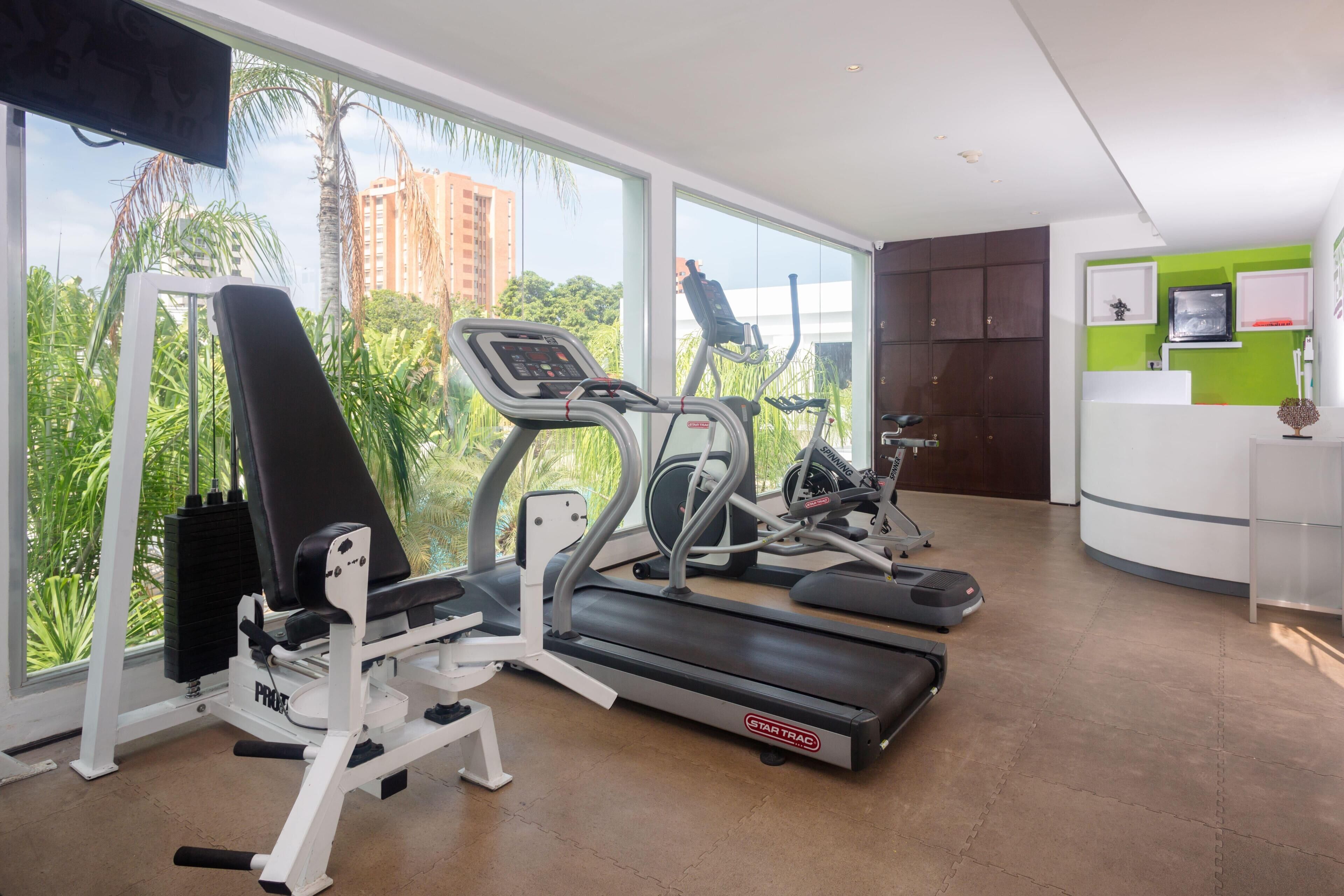 A gym with exercise equipment, large windows, and a view of palm trees and buildings.