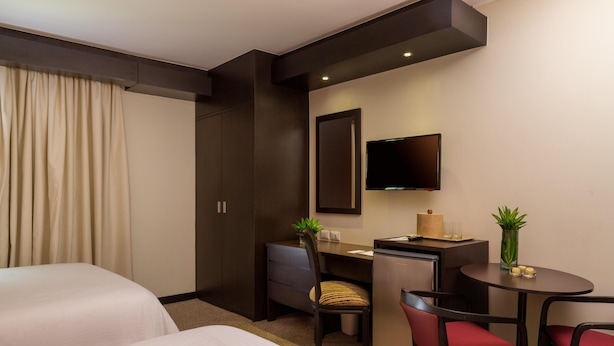 A hotel room with two beds, a desk, a chair, a TV, and a small table with a plant. - Hotel Kristoff (Maracaibo)