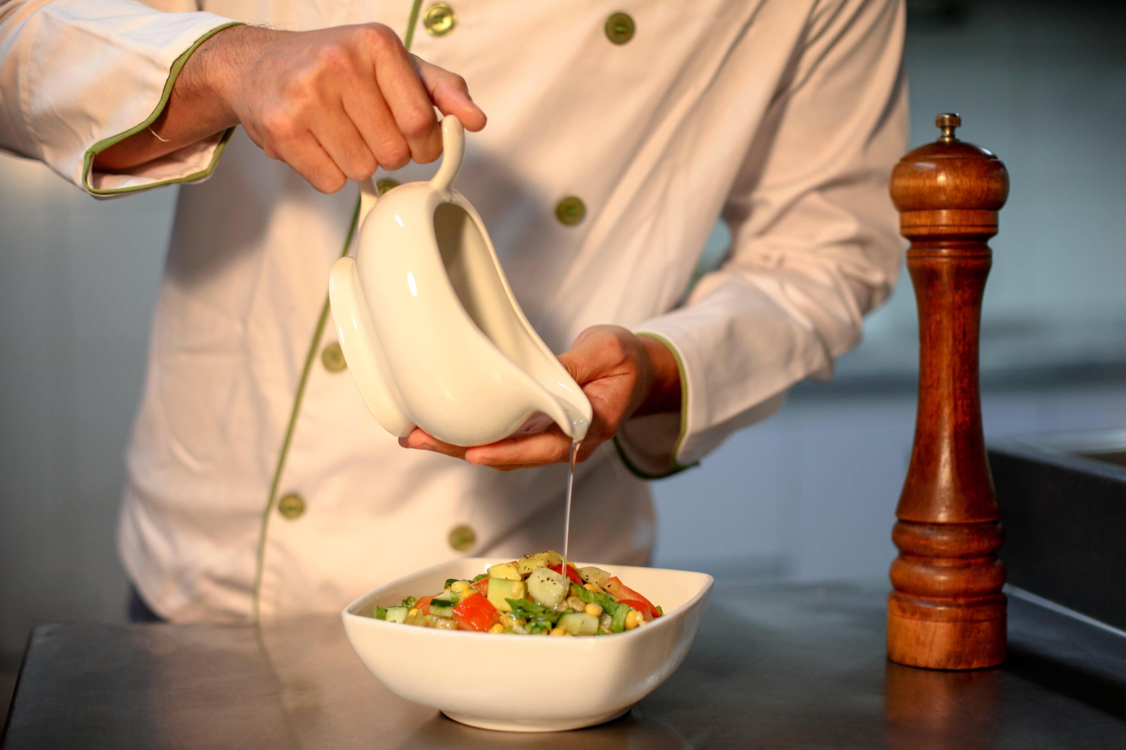 Uma pessoa com avental de chef despejando um líquido branco de um jarro sobre uma salada.