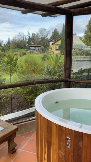 Un bain à remous en bois installé sur une terrasse carrelée, avec vue sur une maison et la verdure environnante.