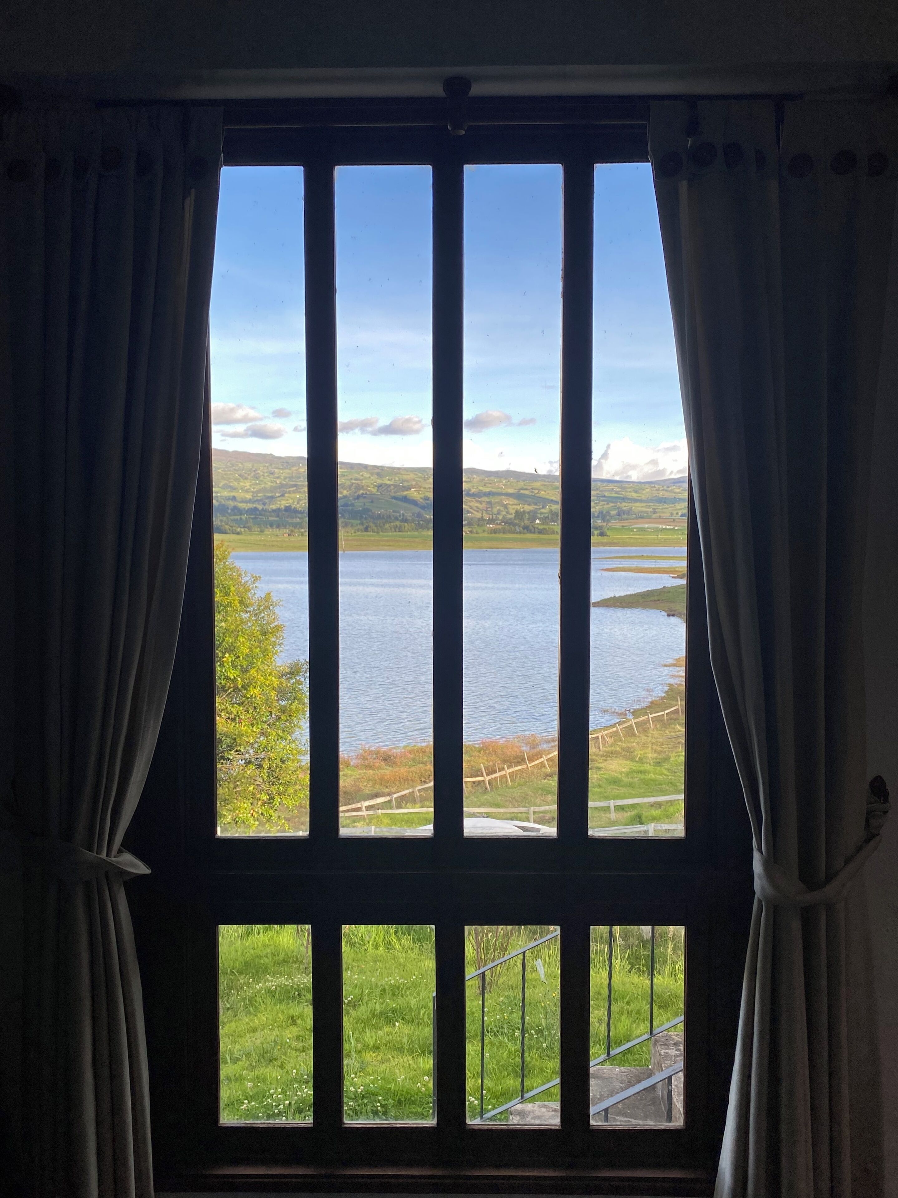 Una ventana con cortinas, vista a un lago y una zona ajardinada con valla.