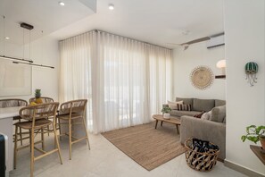 A modern living room with a sofa, a dining area with chairs, a coffee table, and a large window with sheer curtains. - Ground-floor 1BR at Mareal • Walk to the Beach (Cabarete)