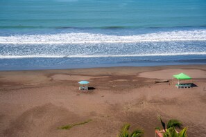 A beach with two umbrellas and beach chairs, a green tent, and a palm tree. - Diamante 701n - Downtown Jaco Deluxe Penthouse (Jaco)