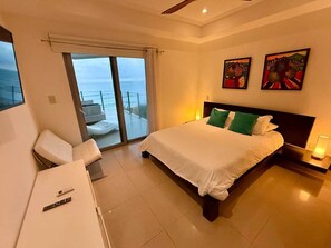 A hotel room with a bed, a desk, a chair, a balcony with a view of the sea, and artwork on the wall. - Diamante 701n - Downtown Jaco Deluxe Penthouse (Jaco)