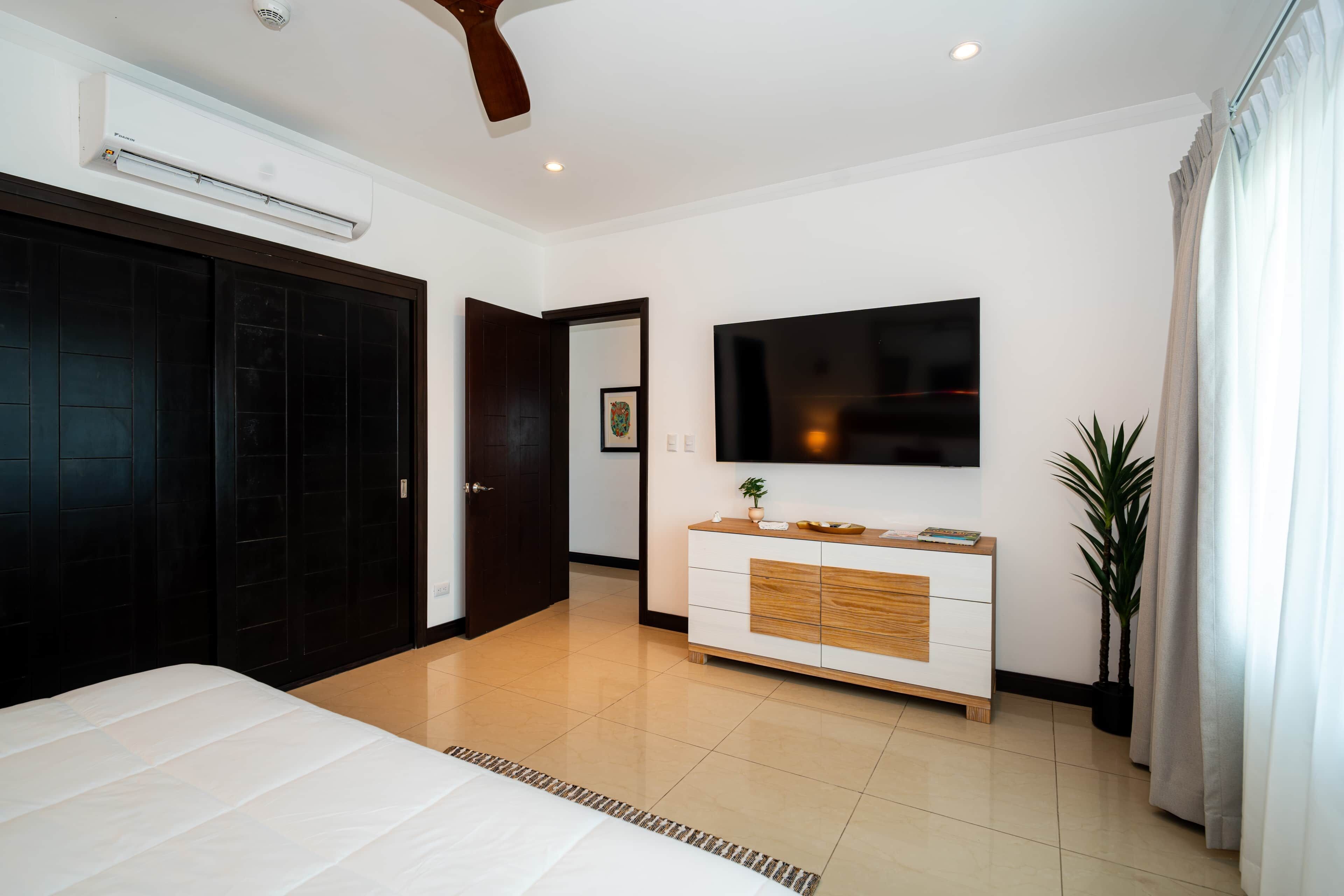 A modern bedroom with a large bed, a wardrobe, a TV mounted on the wall, and a potted plant.