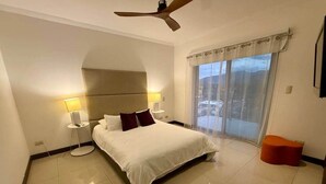 Ein Hotelzimmer mit Bett, Nachttisch, Lampe, Deckenventilator, einem Fenster mit Vorhängen und einem kleinen orangefarbenen Hocker.
