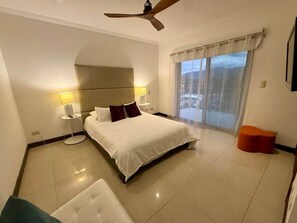 A hotel room with a bed, a nightstand, a lamp, a ceiling fan, a window with curtains, and a small orange ottoman. - Diamante 701n - Downtown Jaco Deluxe Penthouse (Jaco)