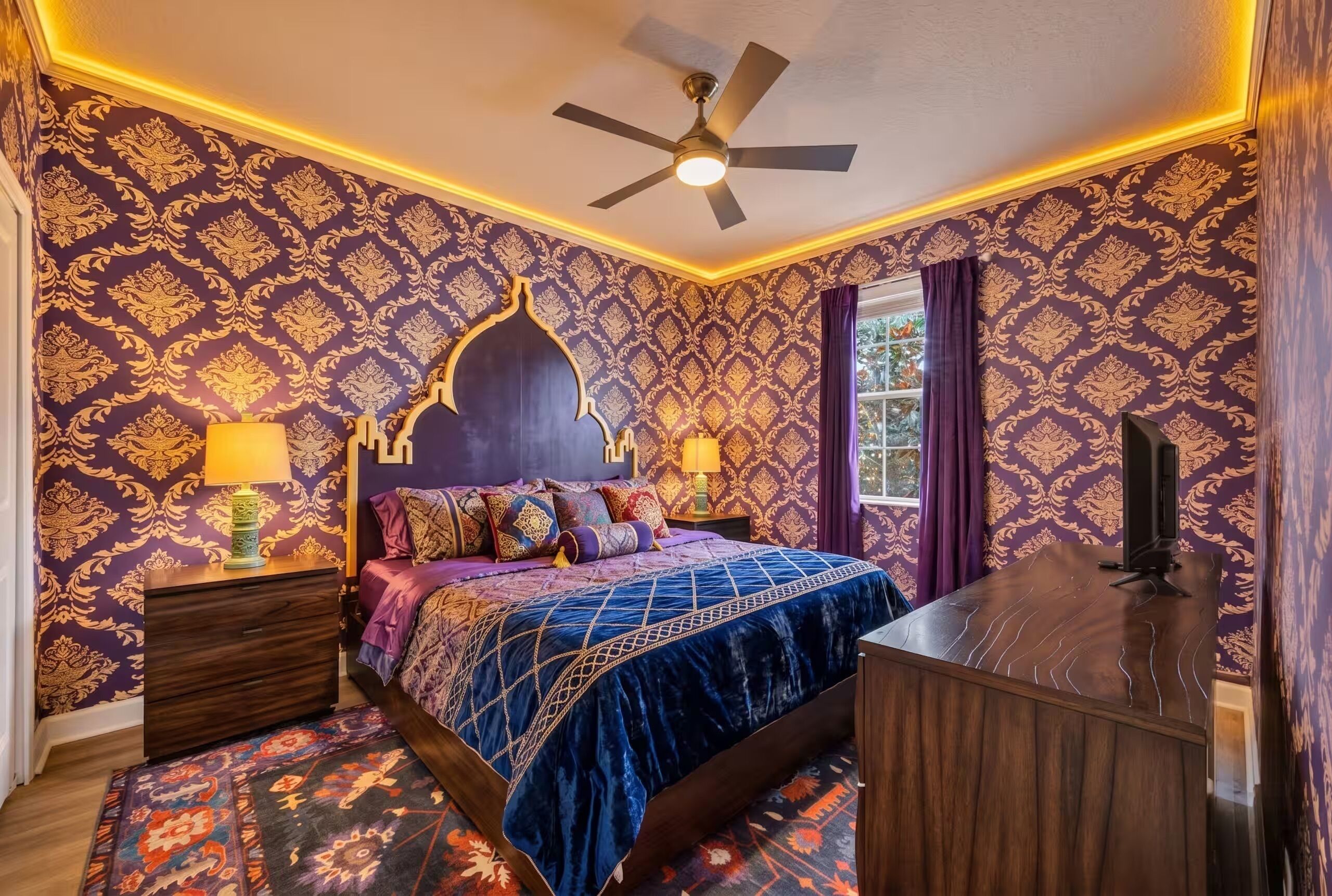A bedroom with a bed, nightstands, a desk, a ceiling fan, patterned wallpaper, and a window with curtains.