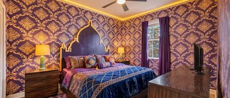 A bedroom with a bed, nightstands, a desk, a ceiling fan, patterned wallpaper, and a window with curtains.
