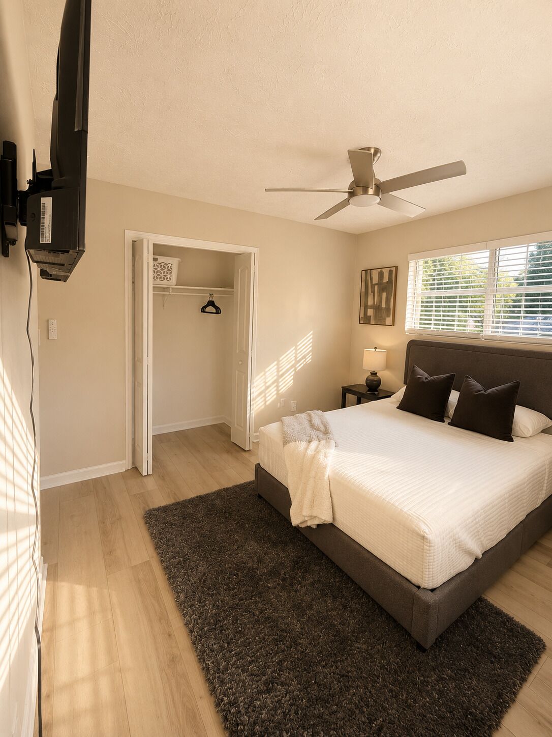 A bedroom with a bed, a ceiling fan, a nightstand, a lamp, a closet, and a window with blinds.