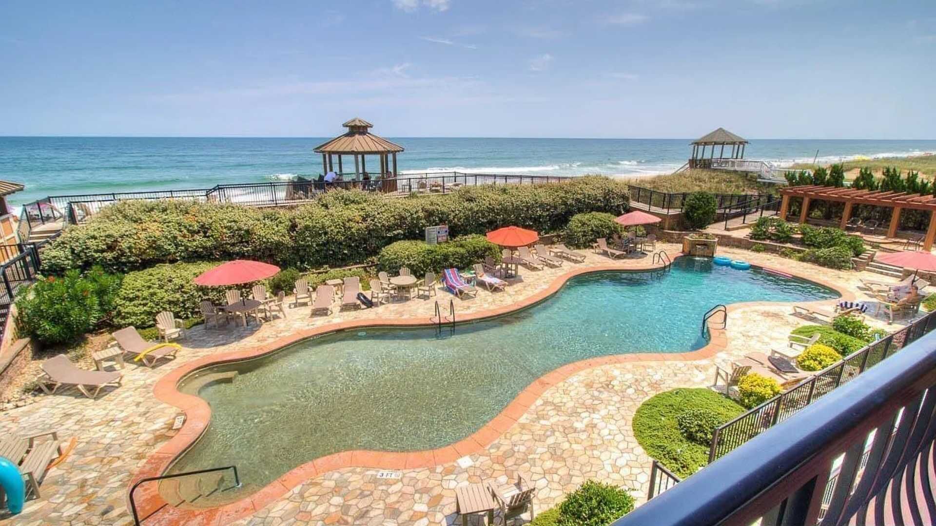 Zona de piscina con sillas de descanso, sombrillas y gazebos con vistas al océano.